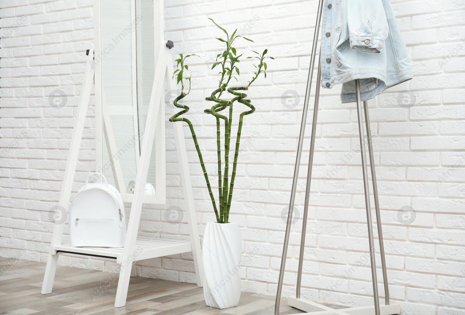 Photo of Vase with green bamboo stems, mirror and rack on floor near white brick wall in room. Interior design