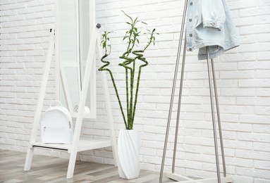 Vase with green bamboo stems, mirror and rack on floor near white brick wall in room. Interior design