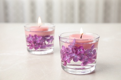 Glass holders with water, lilac flowers and burning candles on light table, space for text