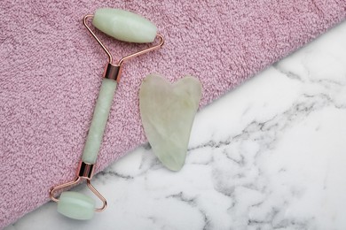 Photo of Gua sha stone, face roller and towel on white marble table, flat lay. Space for text