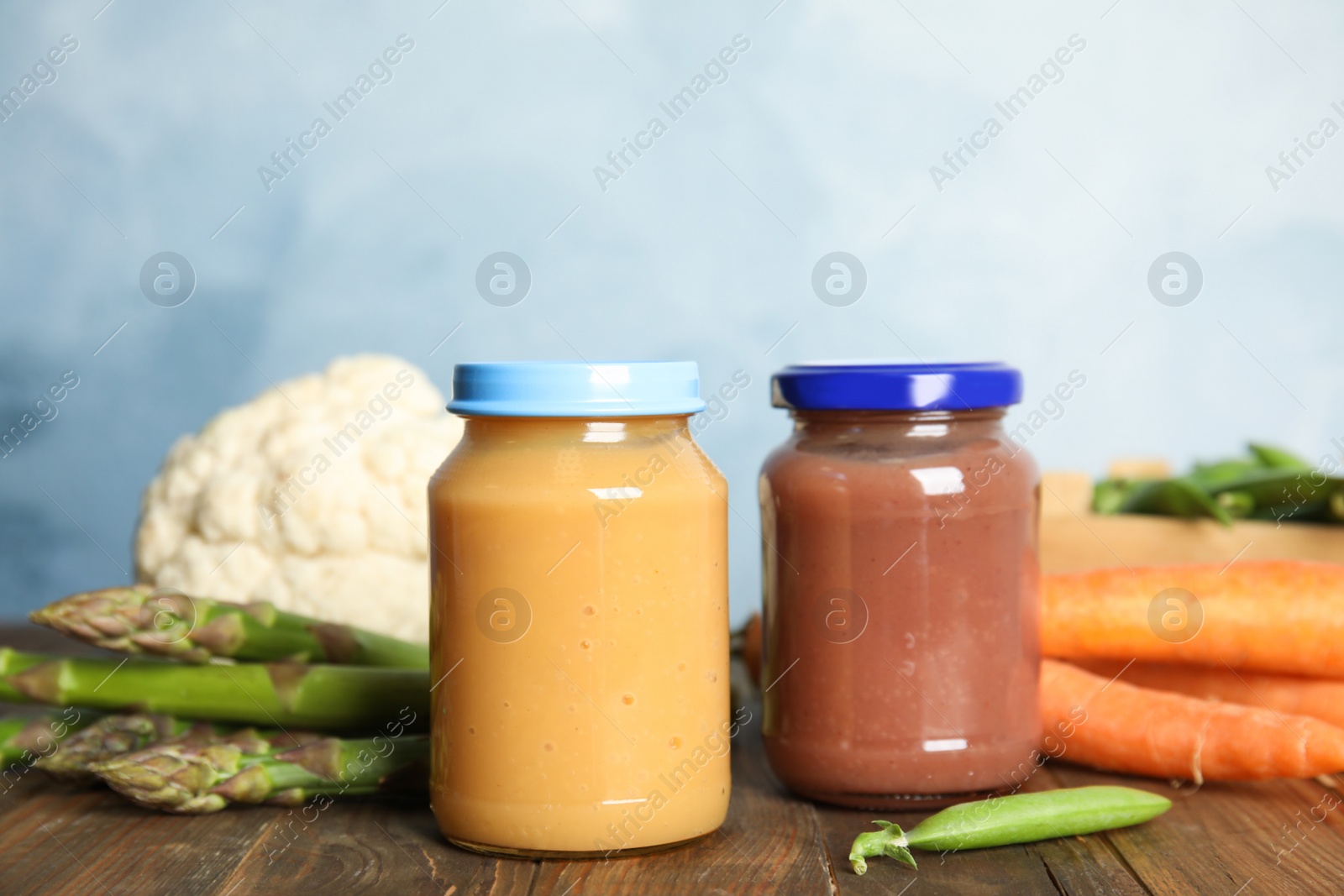 Photo of Jars with baby food and fresh vegetables on wooden table against light blue background