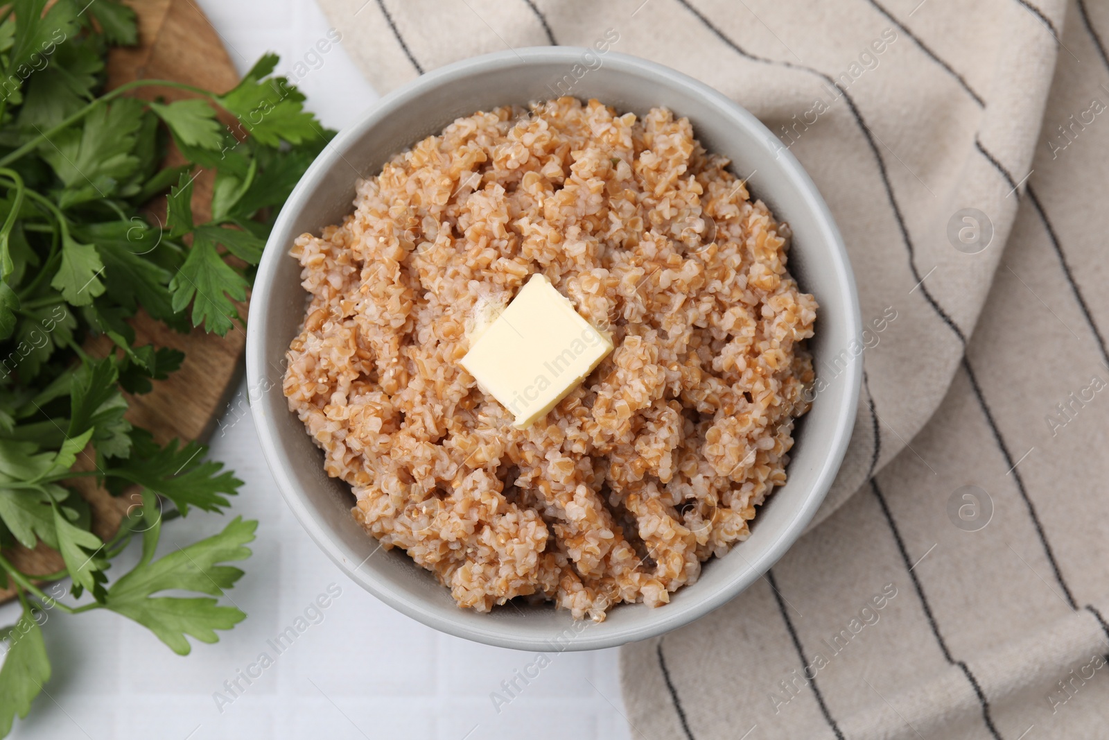 Photo of Tasty wheat porridge with parsley and butter on white tiled table, top view