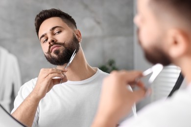 Photo of Handsome young man trimming beard with scissors near mirror in bathroom