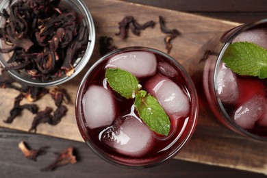 Refreshing hibiscus tea with ice cubes and mint in glasses and roselle petals on wooden table, flat lay