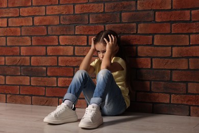 Photo of Child abuse. Upset little girl sitting on floor near brick wall indoors