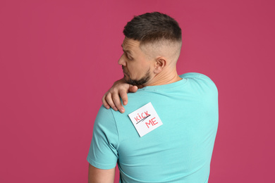 Photo of Man with KICK ME sticker on back against pink background. April fool's day