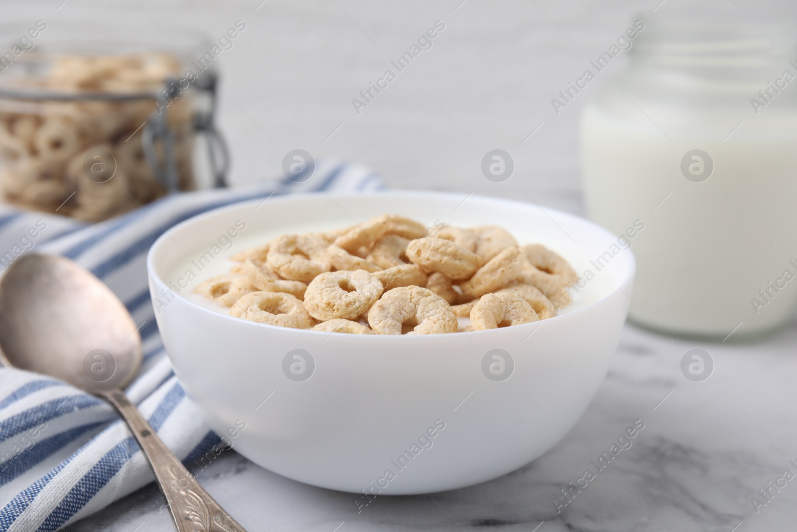 Photo of Breakfast cereal. Tasty corn rings with milk in bowl and spoon on white marble table, closeup