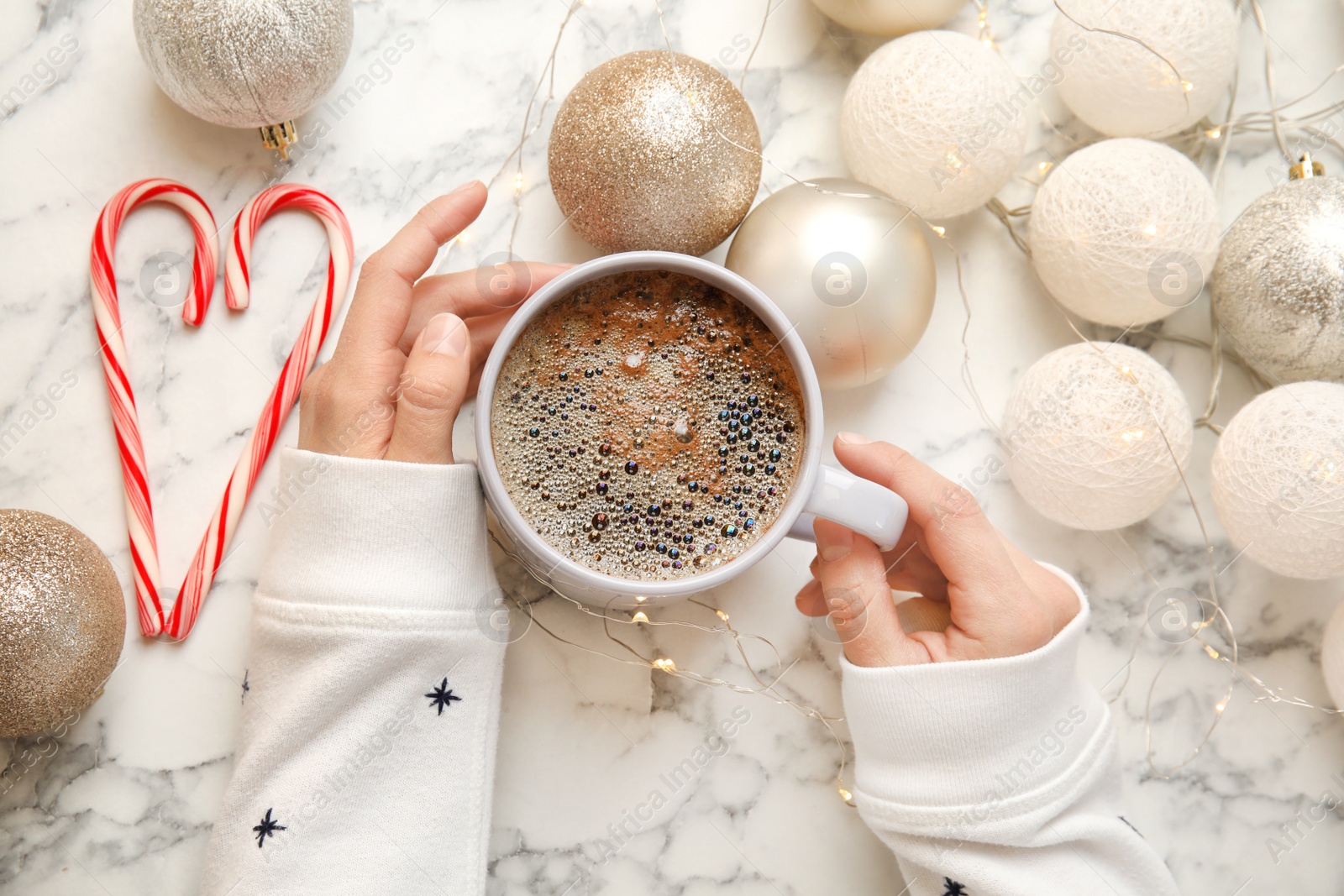 Photo of Woman holding cup of hot winter drink near Christmas lights, balls and candy canes on marble background, top view