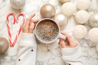 Photo of Woman holding cup of hot winter drink near Christmas lights, balls and candy canes on marble background, top view