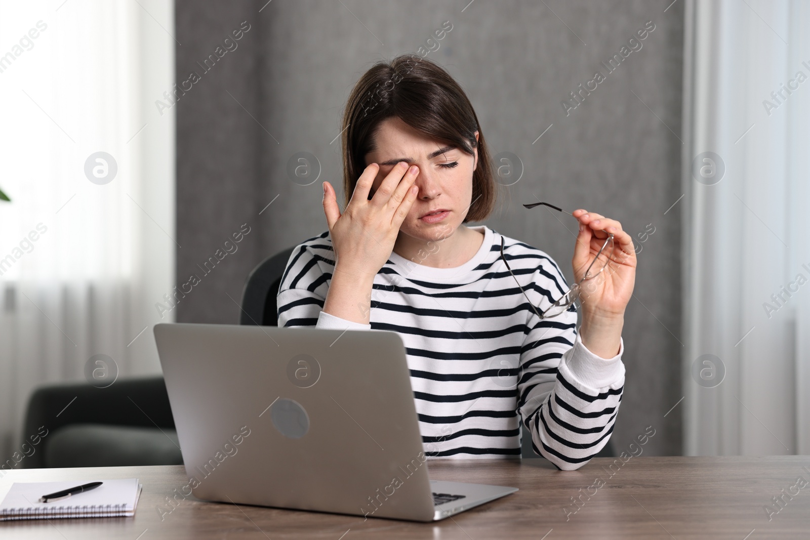 Photo of Overwhelmed woman holding glasses at wooden table with laptop indoors