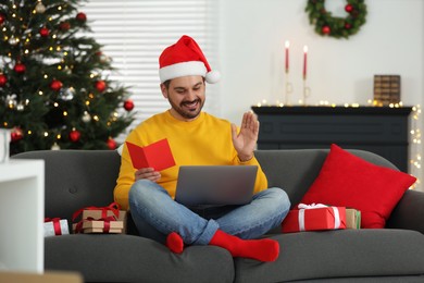 Celebrating Christmas online with exchanged by mail presents. Man with gift and greeting card waving hello during video call on laptop at home