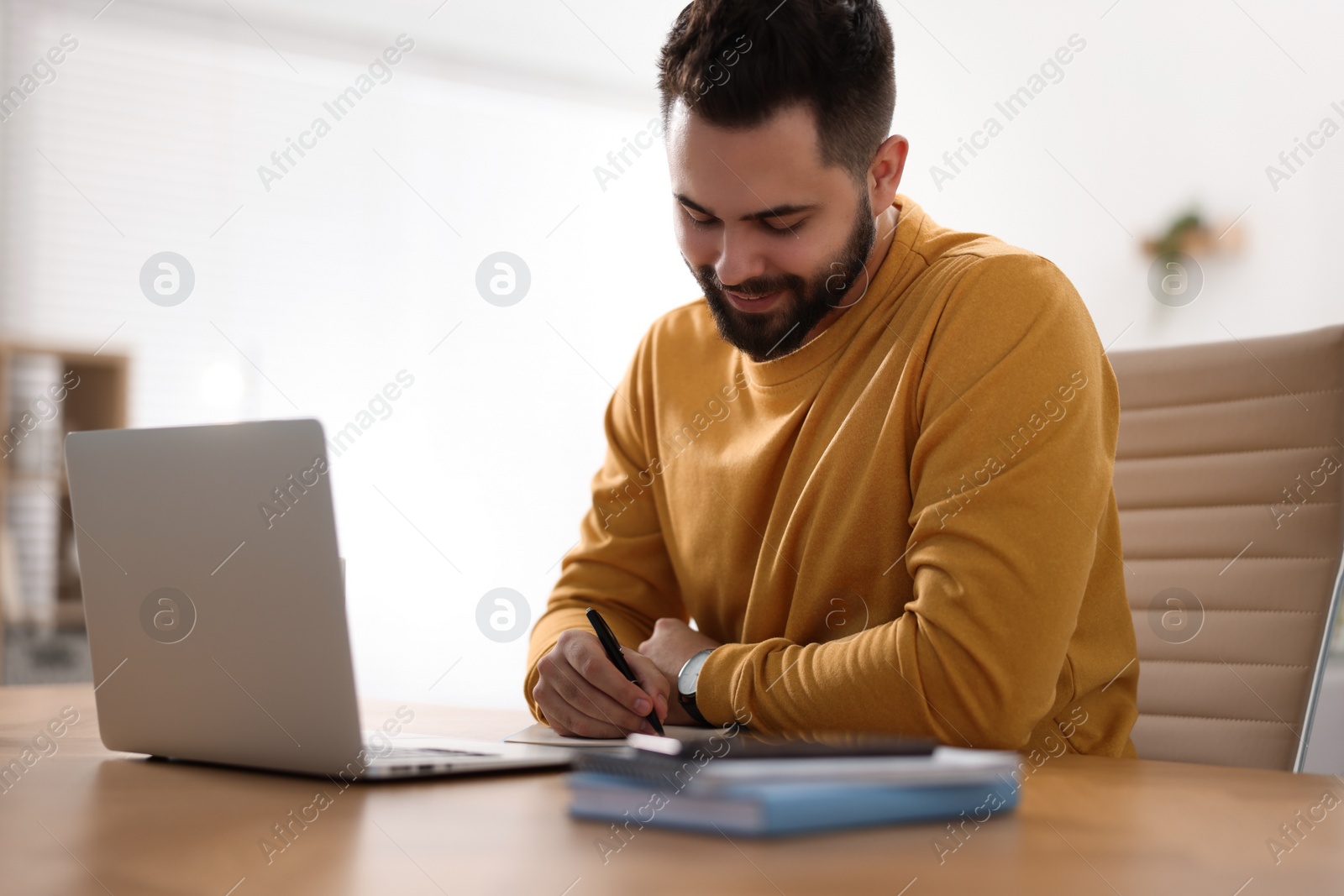 Photo of Young man writing down notes during webinar at table in room