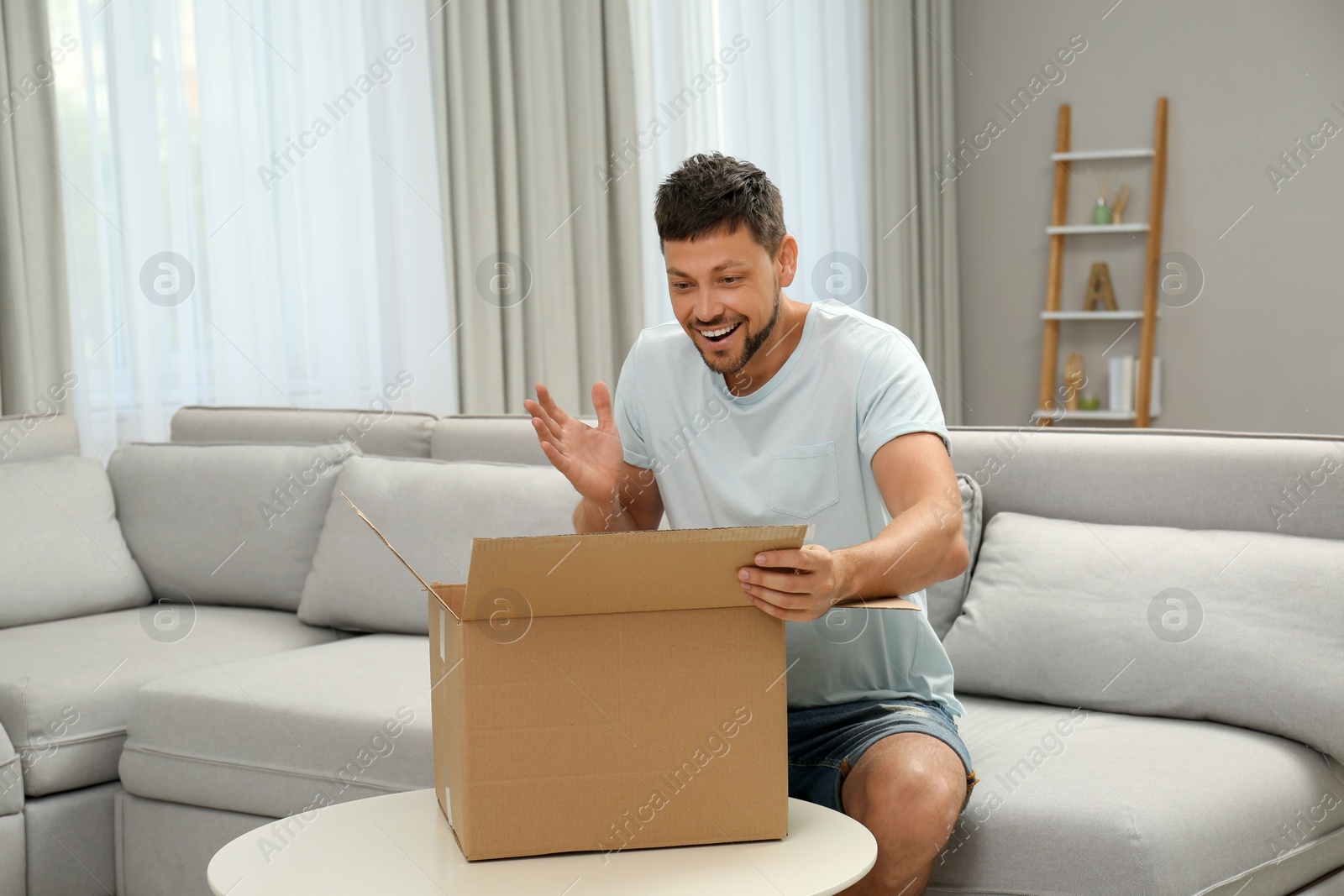Photo of Happy man opening parcel on sofa at home. Internet shopping