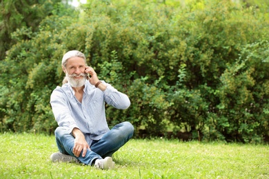 Photo of Handsome mature man with mobile phone sitting on green grass in park