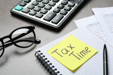 Reminder note with words TAX TIME, documents, glasses and calculator on grey table, closeup