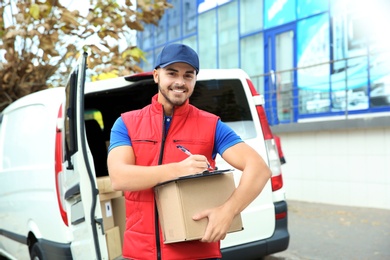 Photo of Young courier with parcel and clipboard near delivery van outdoors