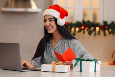 Celebrating Christmas online with exchanged by mail presents. Smiling woman in Santa hat with greeting card and gifts during video call at home