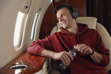 Young man with bottle of water and headphones listening to music in airplane during flight