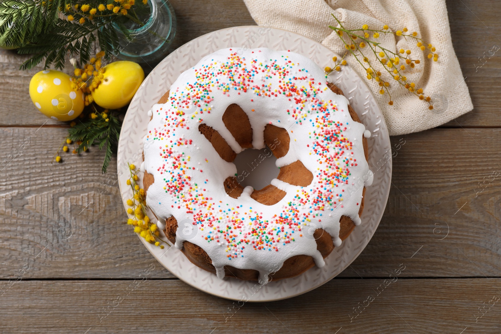Photo of Glazed Easter cake with sprinkles, painted eggs and flowers on wooden table, flat lay