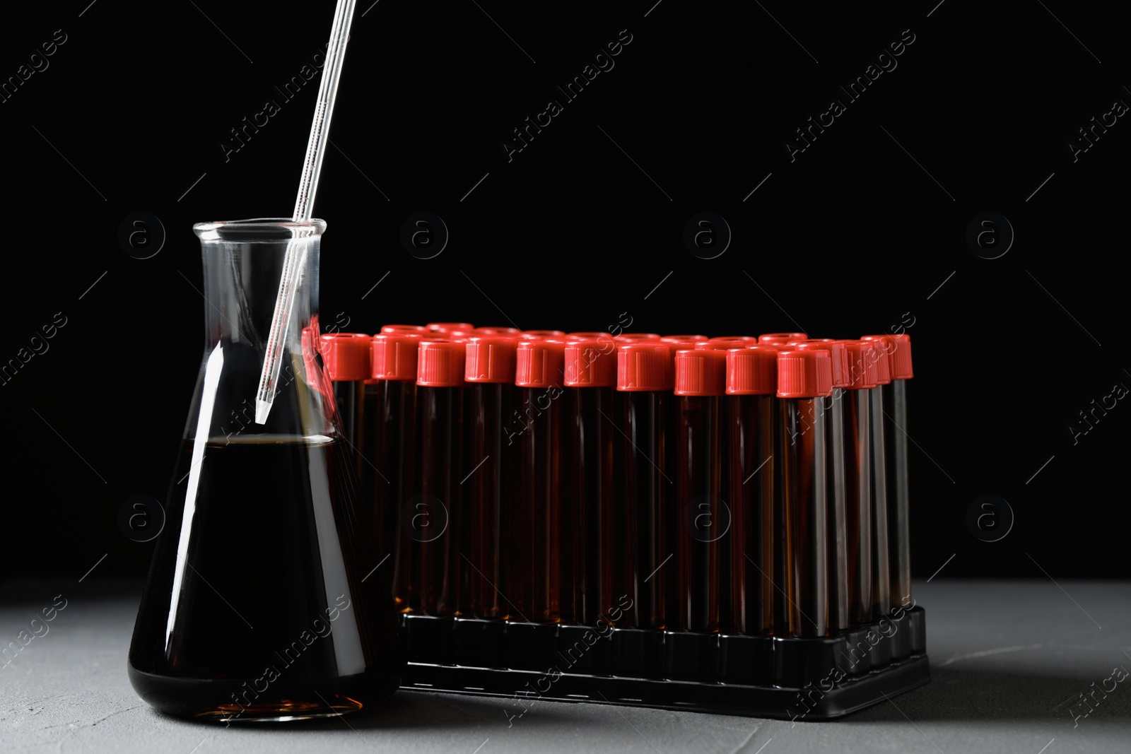 Photo of Different laboratory glassware with brown liquids and pipette on grey table against black background