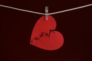 Photo of Broken heart. Torn red paper heart sewed with thread on rope against burgundy background