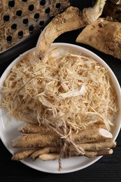 Photo of Plate with whole and grated horseradish roots on black wooden table, flat lay