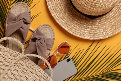 Photo of Wicker bag, smartphone, palm leaves and beach accessories on orange background, flat lay