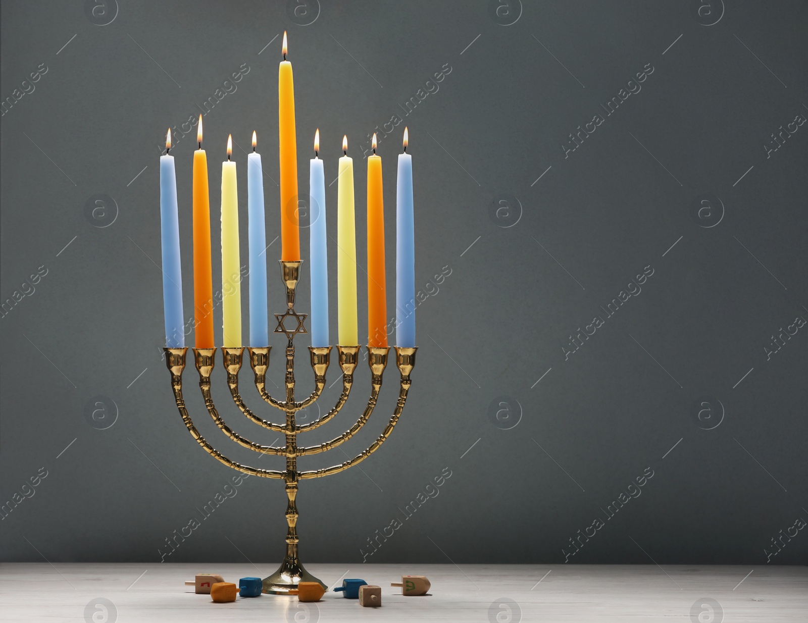 Photo of Hanukkah celebration. Menorah with burning candles and dreidels on light wooden table against grey background, closeup. Space for text