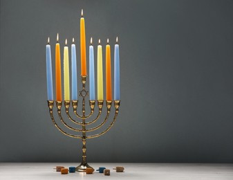 Photo of Hanukkah celebration. Menorah with burning candles and dreidels on light wooden table against grey background, closeup. Space for text