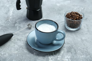 Mini mixer (milk frother), whipped milk in cup and coffee beans on grey textured table