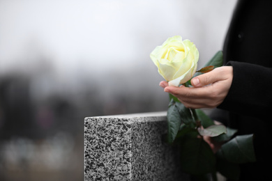 Photo of Woman holding white rose near grey granite tombstone outdoors, closeup. Funeral ceremony