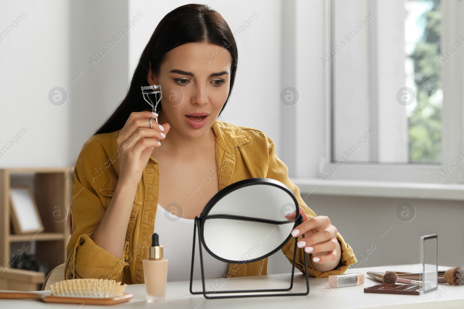 Photo of Emotional young woman using eyelash curler while doing makeup at table indoors