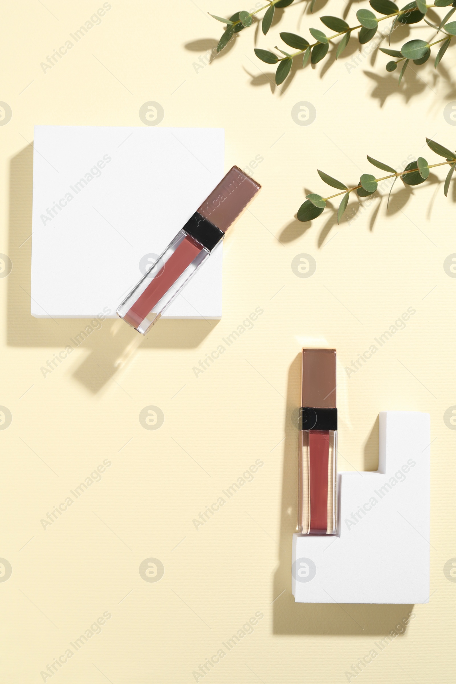 Photo of Lip glosses, podiums and green leaves on pale yellow background, flat lay