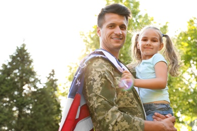 Photo of Father in military uniform with American flag holding his little daughter at green park