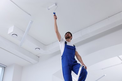 Photo of Handyman with roller painting ceiling on step ladder in room, low angle view