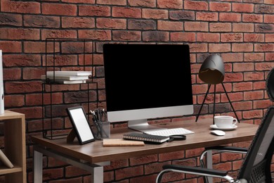 Photo of Cosy workspace with computer, lamp and stationery on desk near brick wall at home