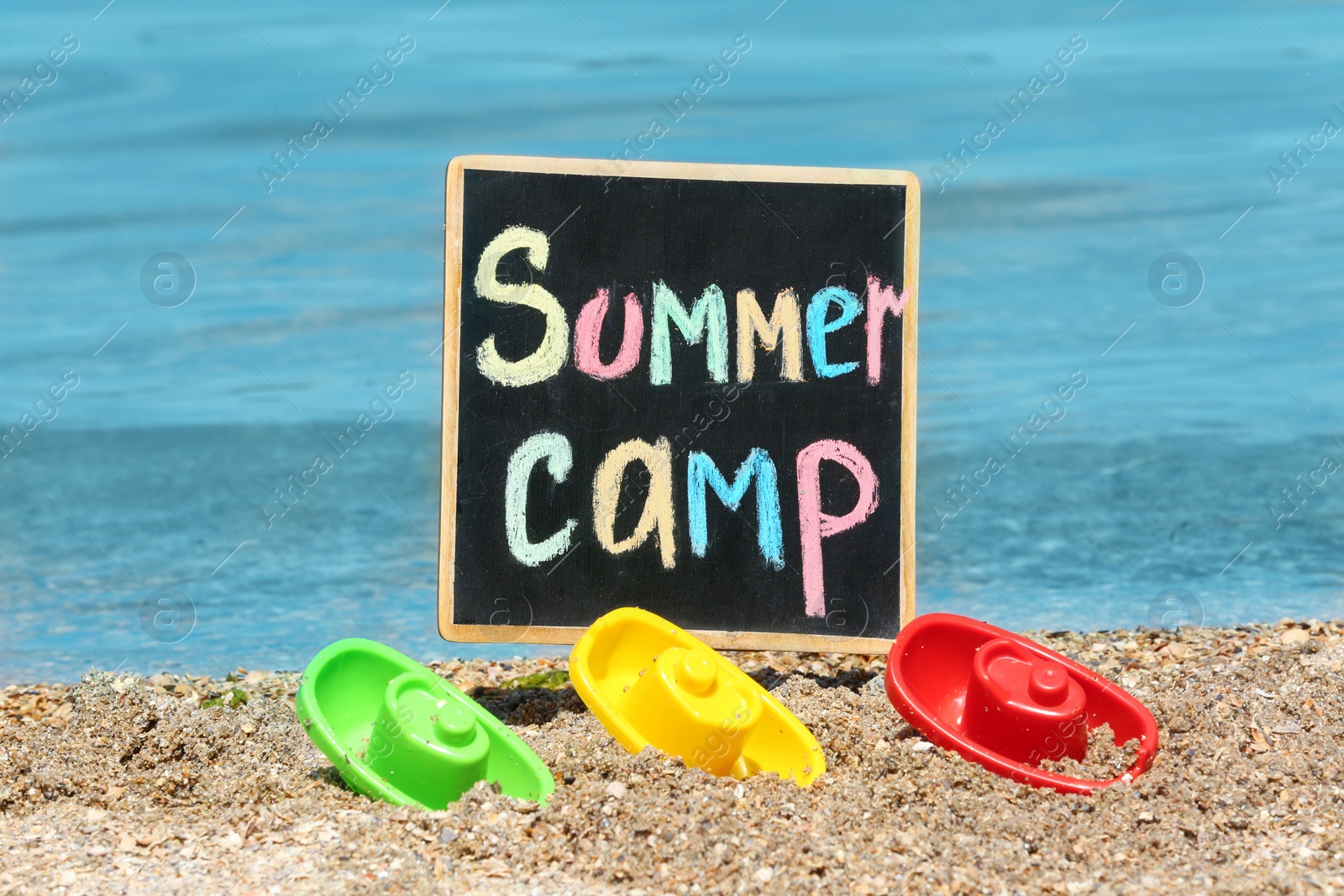 Photo of Little blackboard with text SUMMER CAMP and toys on sand near sea