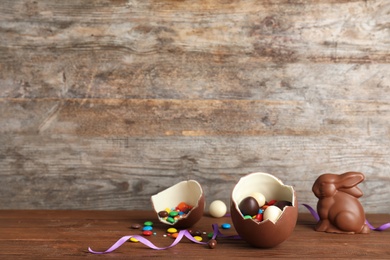 Photo of Broken chocolate Easter egg with candies near bunny on wooden table. Space for text