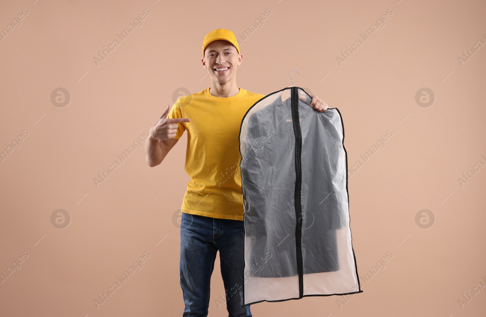 Photo of Dry-cleaning delivery. Happy courier holding garment cover with clothes on beige background