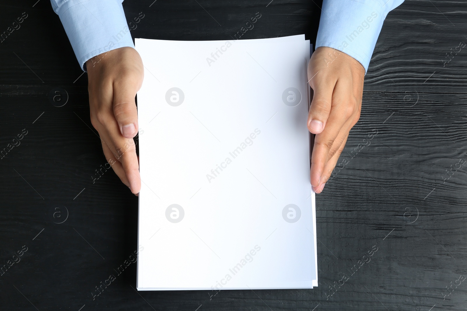 Photo of Man holding blank paper sheets for brochure at black wooden table, top view. Mock up