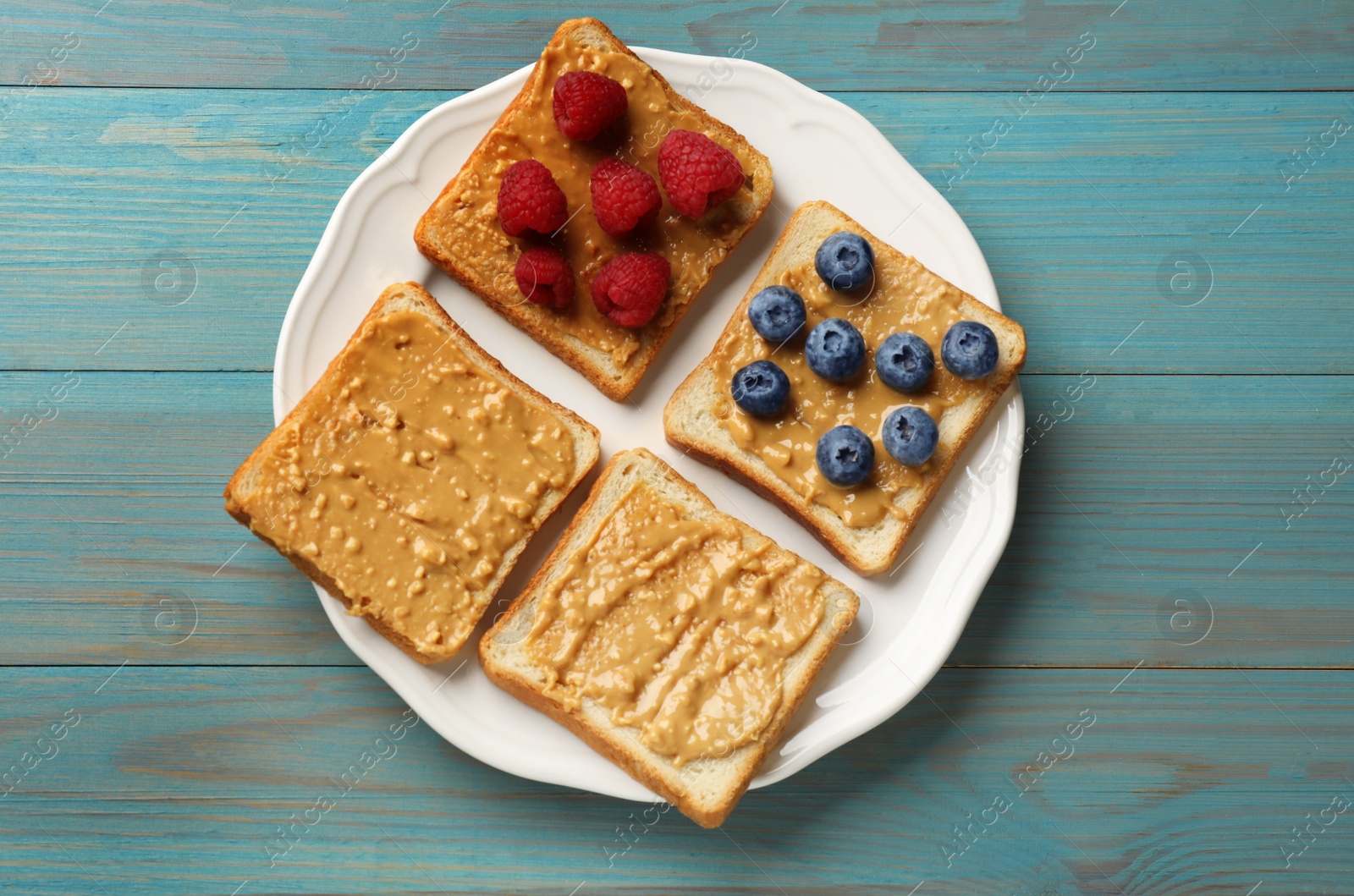 Photo of Delicious toasts with peanut butter, raspberries and blueberries on light blue wooden table, top view