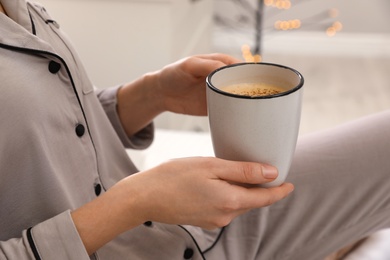 Woman in grey pajama holding cup of coffee indoors, closeup