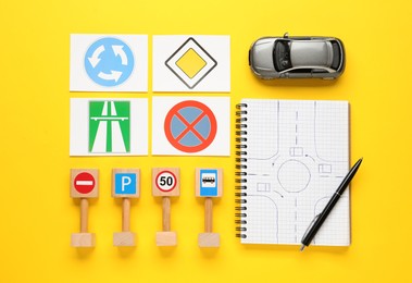 Photo of Many different road signs, notebook with sketch of roundabout and toy car on yellow background, flat lay. Driving school