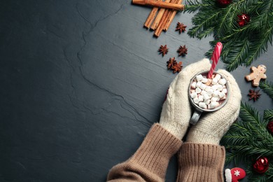 Photo of Woman in knitted mittens holding cup of delicious hot chocolate with marshmallows and candy cane at black table, top view. Space for text
