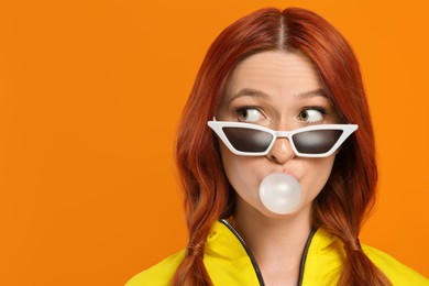Photo of Emotional woman in sunglasses blowing bubble gum on orange background. Space for text