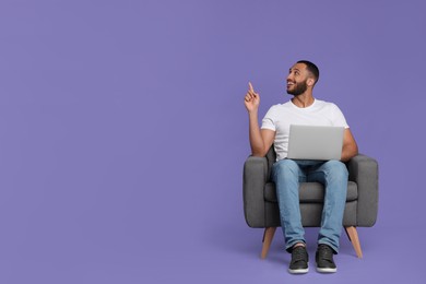 Photo of Smiling young man with laptop sitting in armchair on lilac background. Space for text