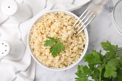 Cooked bulgur with parsley in bowl on white marble table, top view