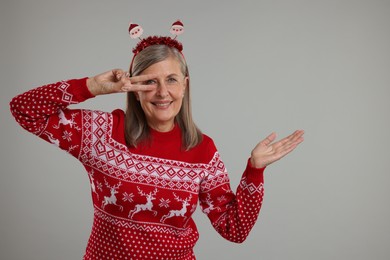 Photo of Happy senior woman in Christmas sweater and Santa headband showing something on grey background. Space for text