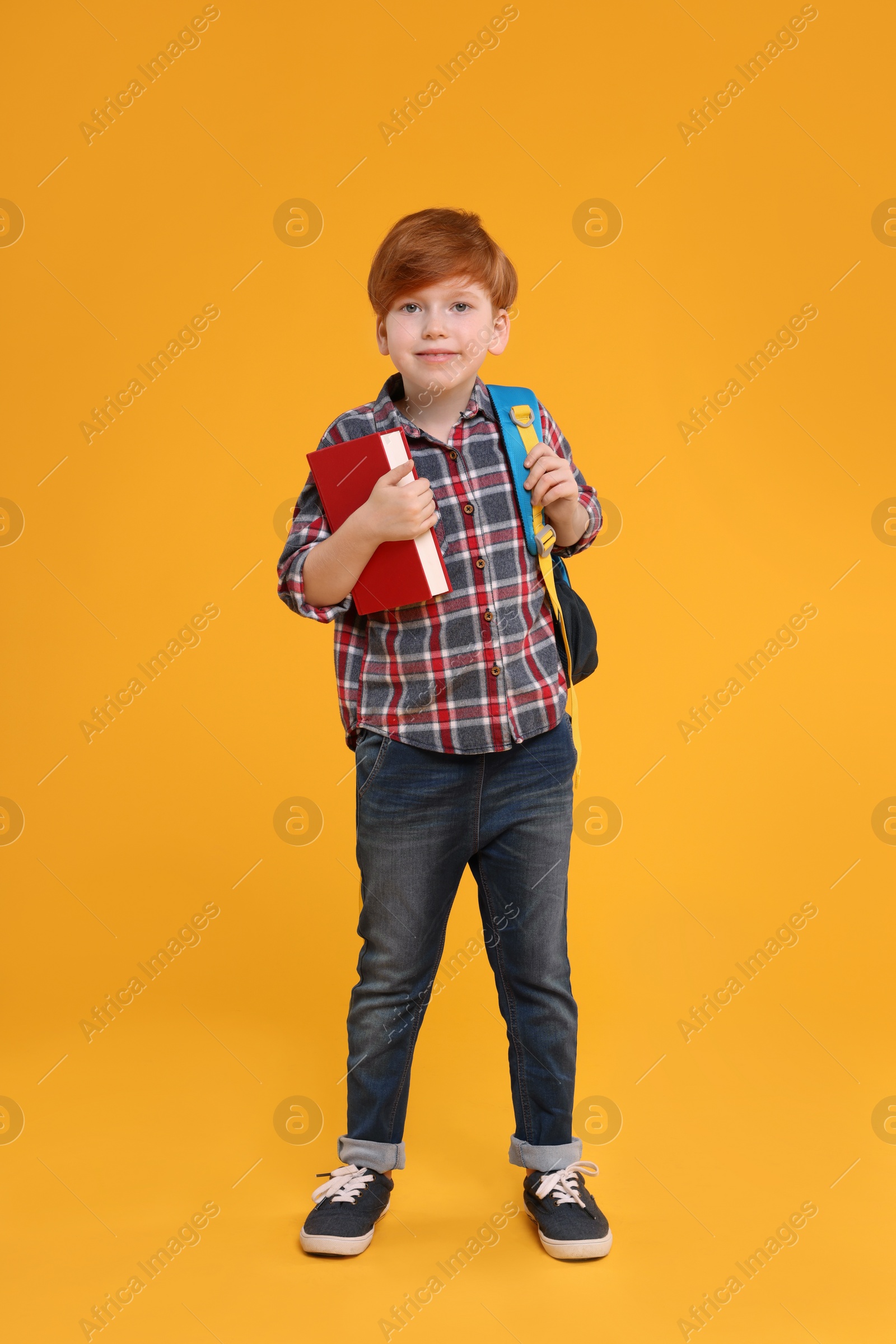 Photo of Cute schoolboy with backpack and book on orange background