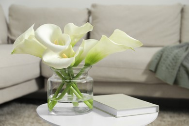 Beautiful calla lily flowers in glass vase and book on white table indoors